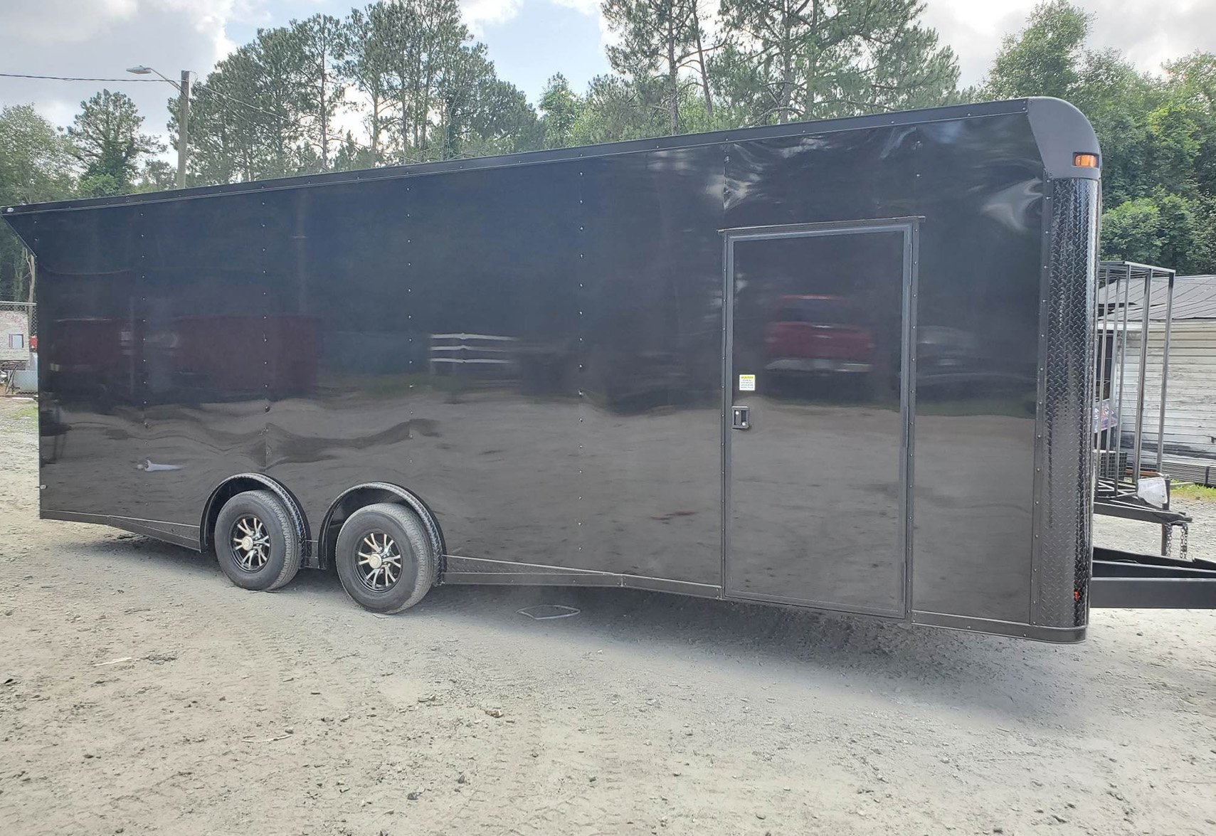 Black enclosed trailer with a flat bed and wheels, parked on a gravel surface.