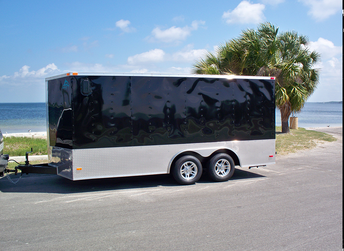 A black enclosed trailer parked on a paved lot with palm trees and a clear blue sky in the background.
